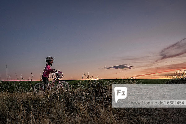 young girl on a bike waits at the top of a grassy hill at sunset