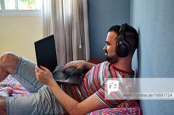 Junger Mann arbeitet mit seinem Laptop in seinem Schlafzimmer  er hört Musik