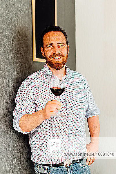 Mann mit Schnurrbart hält ein Glas Rotwein in der Hand und steht an einer Wand