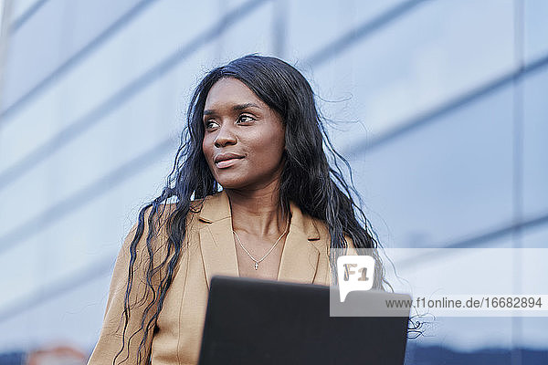 Porträt einer schwarzen Frau in einem braunen Anzug  die auf der Straße an ihrem Laptop arbeitet