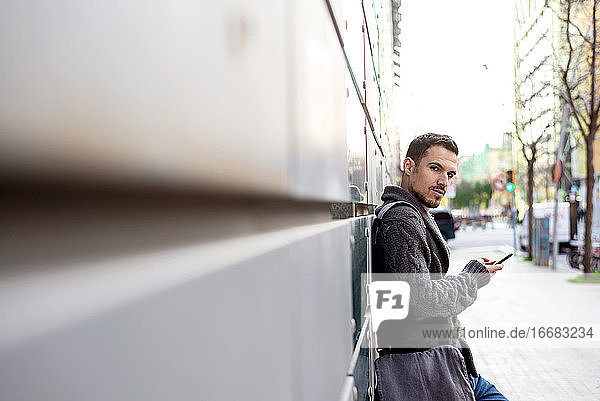 Seitenansicht eines bärtigen Mannes  der ein Telefon benutzt und sich an die Wand eines Bürogebäudes lehnt