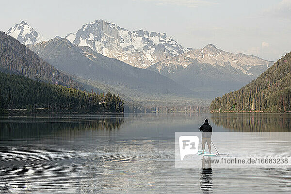 Rückenansicht eines anonymen Mannes auf einem Paddelbrett auf der ruhigen Oberfläche des Duffey-Sees und Genießen der Berglandschaft am Morgen in British Columbia