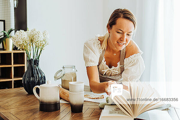 Frau liest ein Buch mit Rezepten  während sie am Küchentisch kocht