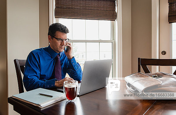 Man on cellphone working from home using a computer at a dining table.