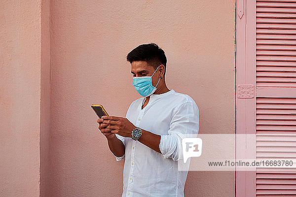 Junger lateinamerikanischer Mann mit Maske schaut auf sein Telefon auf rosa Hintergrund