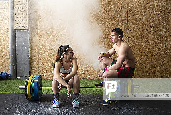 Starker Mann ohne Hemd und Frau in Sportkleidung  die lächeln und sich nett unterhalten  während sie im Fitnessstudio auf einer Langhantel sitzen