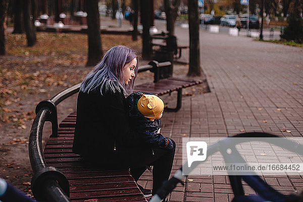 Junge Mutter hält ihr Baby im Arm  während sie auf einer Bank im Park sitzt