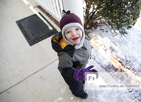 Boy in Winter Gear Standing on Snowy Front Porch Looks Up At Camera