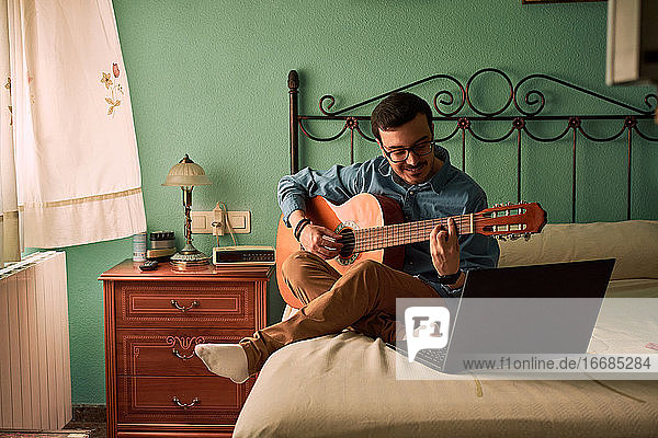 Young man practices with the guitar by looking at his laptop at home