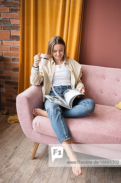 Eine Frau sitzt mit einer Zeitschrift und einer Tasse Tee auf einem rosa Sofa im Wohnzimmer.