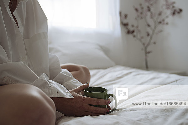 Nahaufnahme von Frauenhänden  die eine Tasse Kaffee auf dem Bett halten.