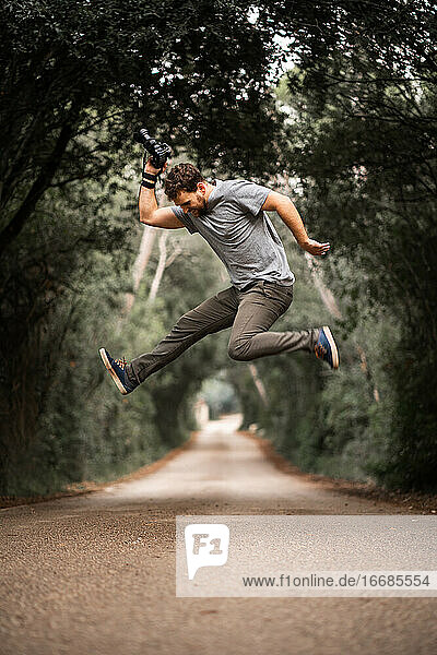 Young photographer jumps on a road where trees make a tunnel