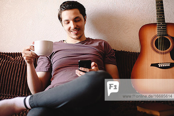 Glücklicher junger Mann  der sein Smartphone benutzt  während er zu Hause Tee trinkt