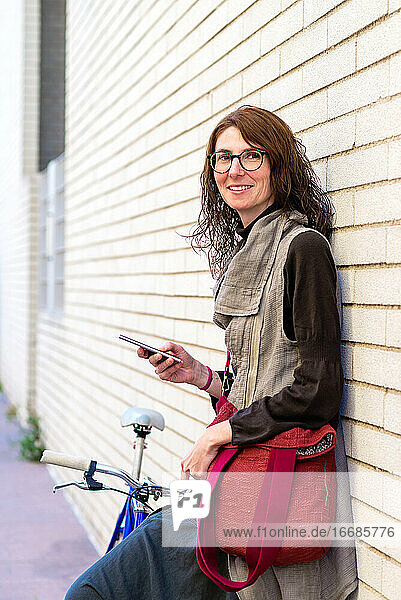 Lächelnde junge Frau mit Fahrrad  die sich an die Wand lehnt und ein Handy benutzt