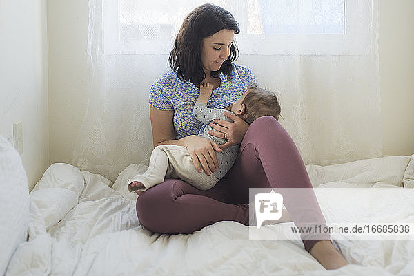 Barefoot mother breastfeeding baby on white bed in front of window