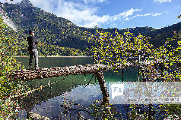 Ein kaukasischer Mann steht auf einem umgestürzten Baum über dem Alpsee in Deutschland