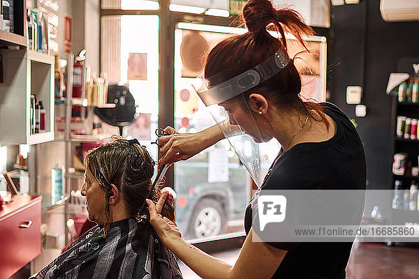 Friseur schneidet einer Kundin die Haare mit einem Gesichtsschutz