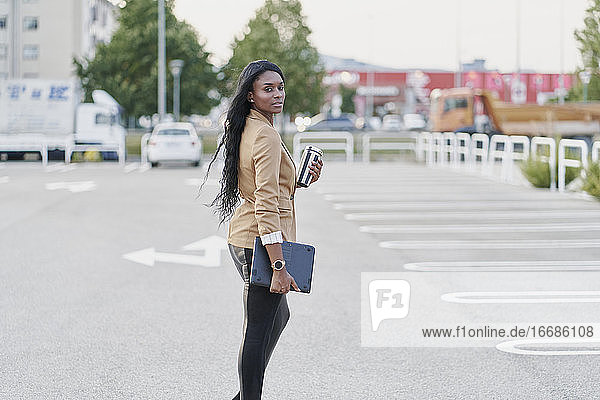 Porträt einer schwarzen Frau in einem braunen Anzug  die einen Laptop und einen Behälter mit Kaffee in der Hand hält und die Straße entlanggeht