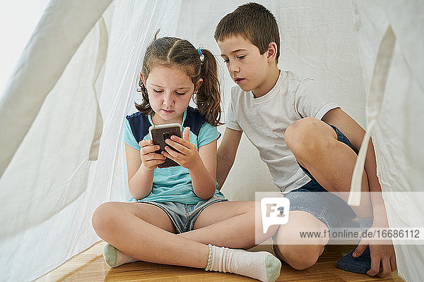 Junge und Mädchen schauen auf ein Smartphone in einem weißen Tipi-Zelt in ihrem Haus. Technologie-Konzept