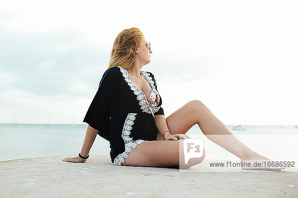 Pensive blonde woman with sunglasses sitting sideways near the sea