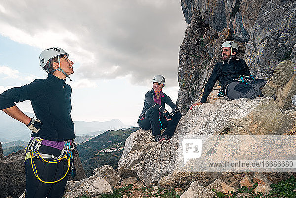 Concept: adventure. Three of climbers with helmet and harness. A man and two women. Resting relaxed sitting on a rock. Via ferrata in the mountains.