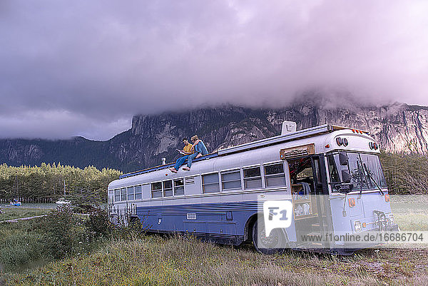 zwei Personen sitzen auf dem Dach eines Schulbusses und lesen ein Buch vor einem Berg