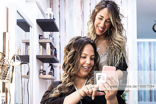 Frau mit Smartphone und Friseurin beim Haarstyling im Friseursalon