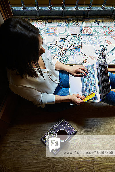 Draufsicht auf eine junge Frau  die mit einer Kreditkarte auf einem Laptop einkauft und online bezahlt  während sie sich zu Hause neben einem Fenster entspannt.