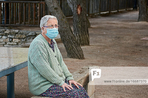 Die Großmutter sitzt auf einer Bank und hat eine Maske auf.