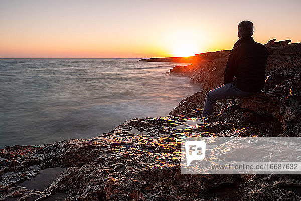 Man sitting on the rocks by the sea watching the sunset. Long exposure