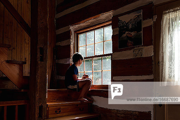 Junge sitzt auf den Stufen eines Blockhauses und schaut durch das Fenster hinaus