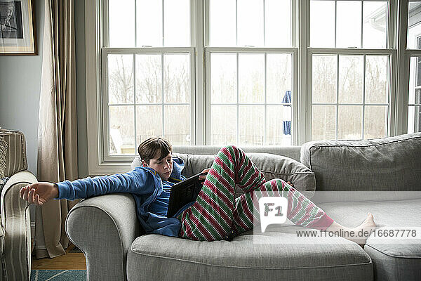 Teenager mit Grippe  im gestreiften Pyjama  sitzt auf der Couch und schaut auf das Ipad