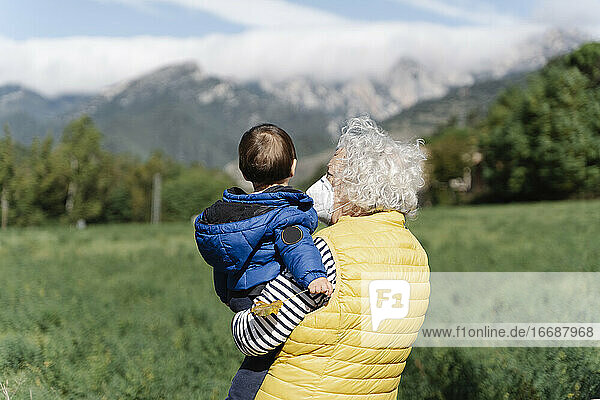 Rückansicht einer Großmutter mit einer medizinischen Maske  die ihren kleinen Enkel auf einem Feld trägt