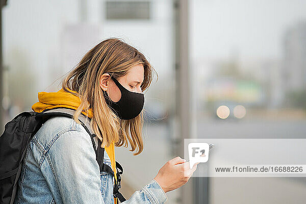 Frau mit Maske und Mobiltelefon an einer Bushaltestelle. Junge Frau benutzt