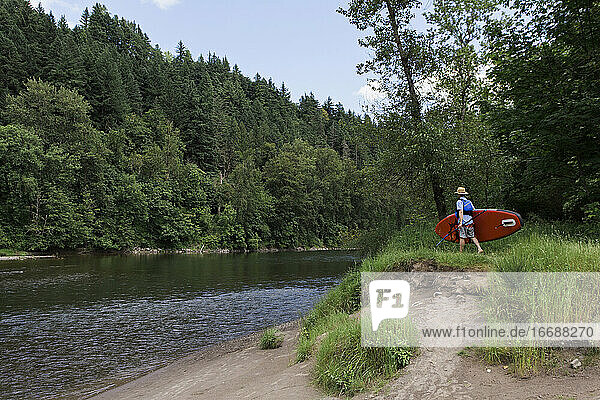 A young man carries his standup paddleboard to a river in Oregon.