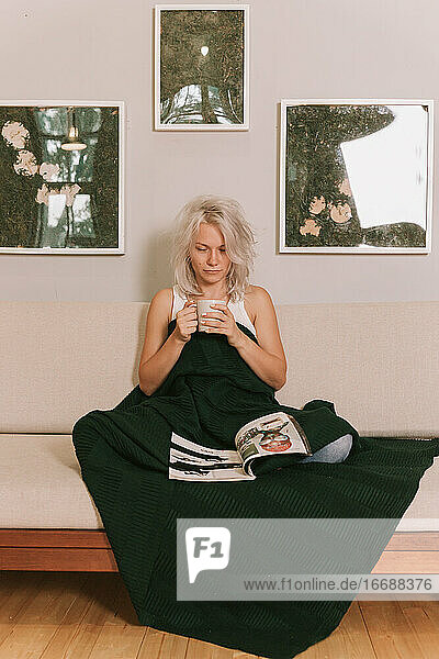Junge Frau sitzt auf einem Sofa unter einem Plaid und trinkt Kaffee