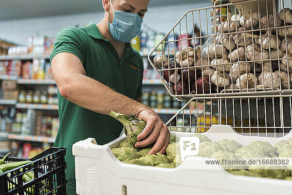 Ein Ladenbesitzer mit einer Maske  der Artischocken aus einer Kiste im Obstladen nimmt.