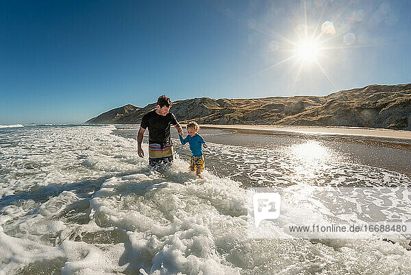 Lächelnder Vater und Kind spielen in den Wellen an einem sonnigen Tag an einem Strand in Neuseeland
