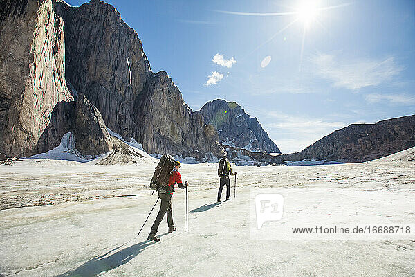 Two backpackers hiking on glacier below steep mountains.