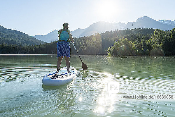 Rückenansicht einer barfuß lebenden Frau in Kleid und Schwimmweste auf einem SUP-Board auf einem ruhigen See an einem sonnigen Sommertag in den Bergen in British Columbia  Kanada