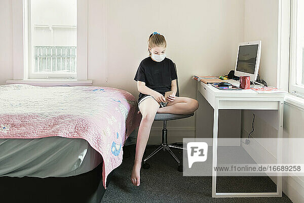 Junges Mädchen mit Gesichtsmaske schaut auf ein Gerät in ihrem Schlafzimmer