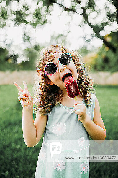 Junges Mädchen isst ein Eis am Stiel und gibt draußen ein Friedenszeichen