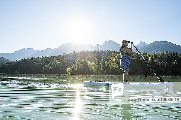 Seitenansicht einer schwangeren Frau in Schwimmweste auf einem SUP-Board auf einem ruhigen See vor einem Bergkamm und Wald an einem sonnigen Tag in British Columbia  Kanada