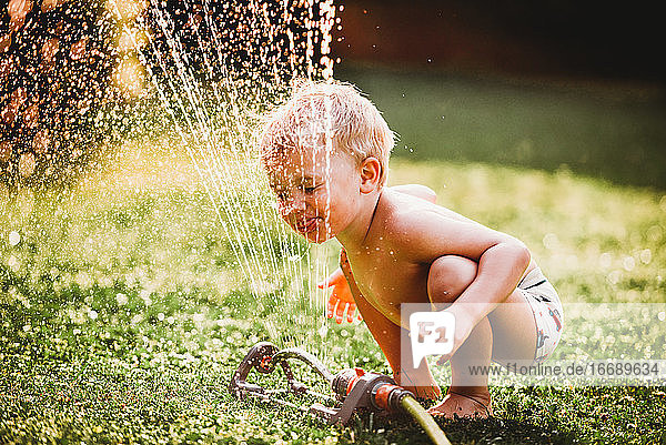 Weißer Junge streckt die Zunge heraus und trinkt Wasser aus dem Rasensprenger im Garten