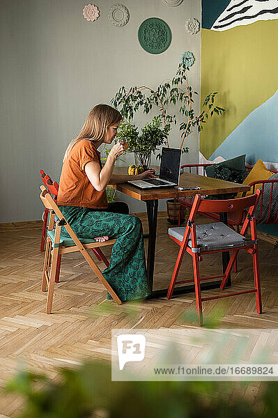 Frau mit Glastasse Tee arbeitet online zu Hause am Laptop auf dem Tisch
