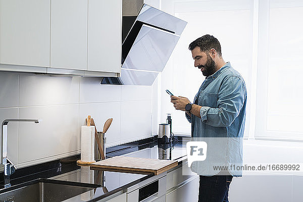 Mann kocht Crêpes in der Küche mit einem Mobiltelefon in einem Jeanshemd