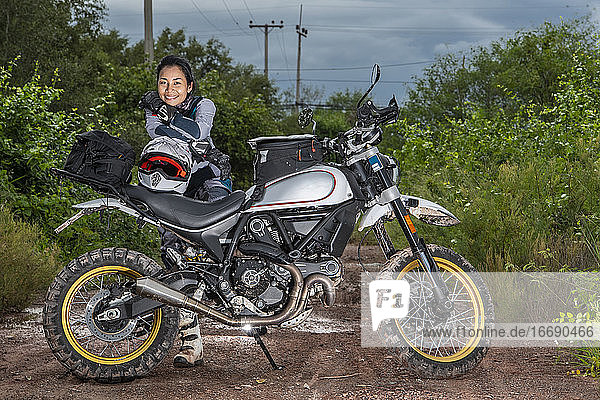 Frau posiert hinter ihrem Scrambler-Motorrad auf einem unbefestigten Weg