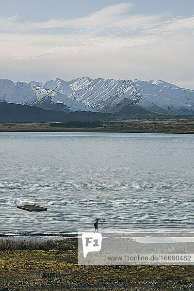 Young man walks next to lake Tekapo with mountains in the background.