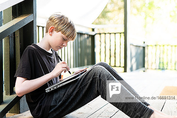 Junge Person sitzt auf einer Veranda und benutzt ein Tablet und einen Stift