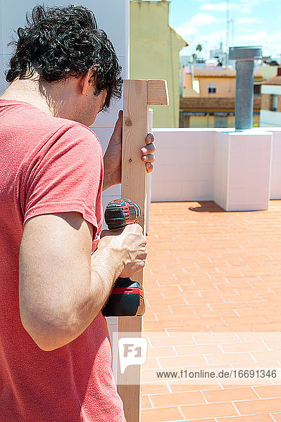 Ein Mann baut an einem sonnigen Tag mit einer Bohrmaschine ein Regal auf einem Dach.